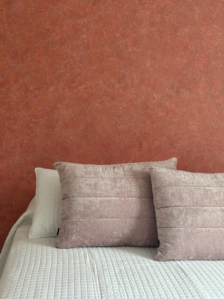 Una cama con cubrecolcha blanco acolchado y dos almohadas, una clara y otra rosa, frente a una pared roja con textura.