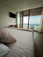 Un dormitorio con una cama grande, un televisor montado en la pared y vista al océano a través de amplias ventanas.