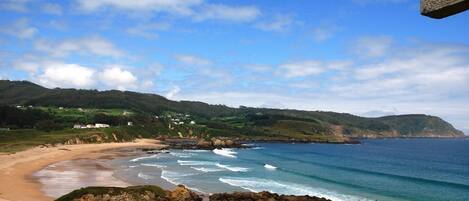 A coastal landscape with a sandy beach, rocky shoreline, and green hills in the background.