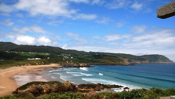 Un paisaje costero con una playa arenosa, costa rocosa y colinas verdes al fondo.