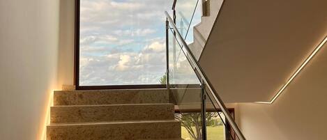 Una escalera moderna con barandillas de cristal y vista al cielo a través de un ventanal grande.