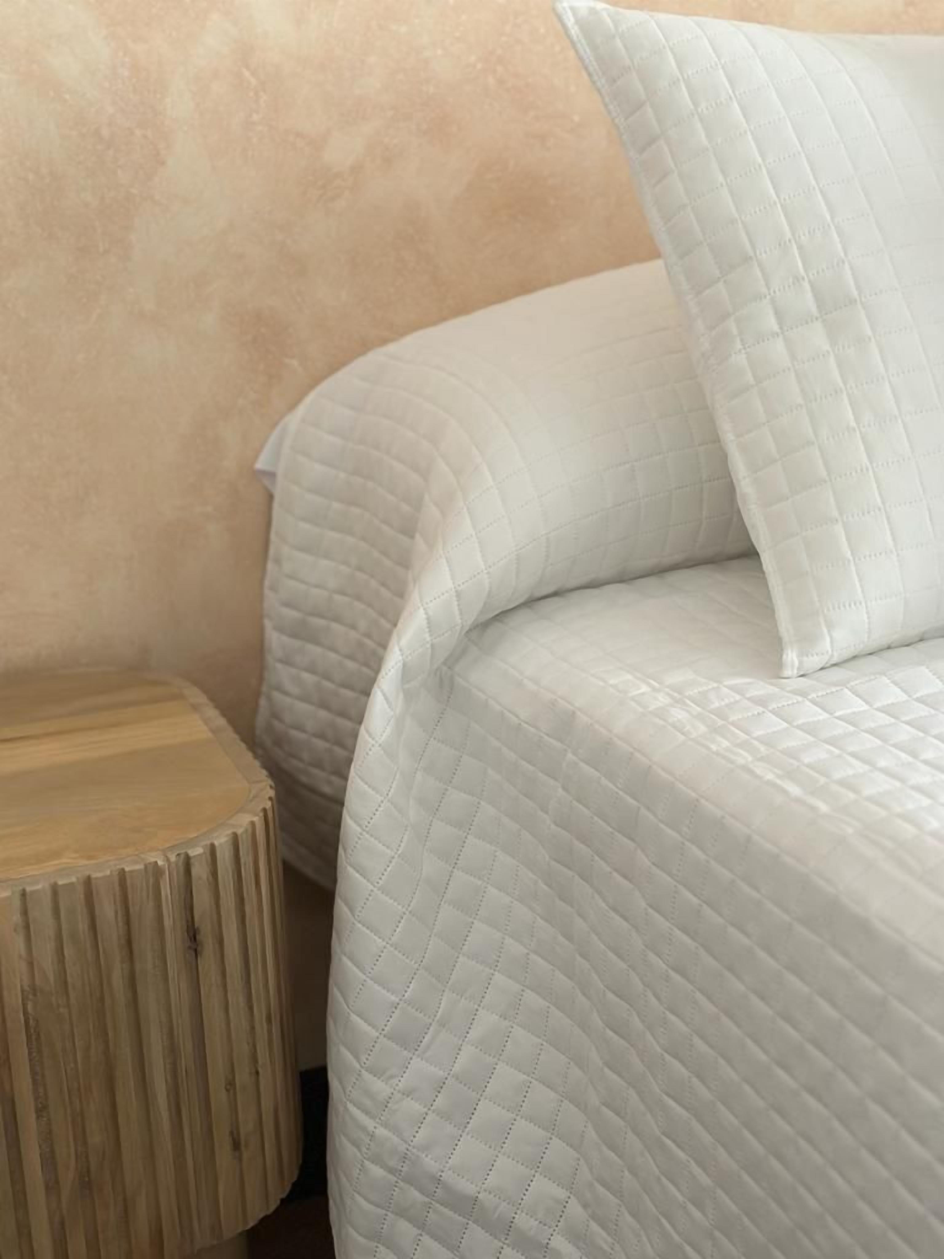 A bed with a quilted mattress and pillows, a wooden nightstand beside it, and a textured wall in the background.