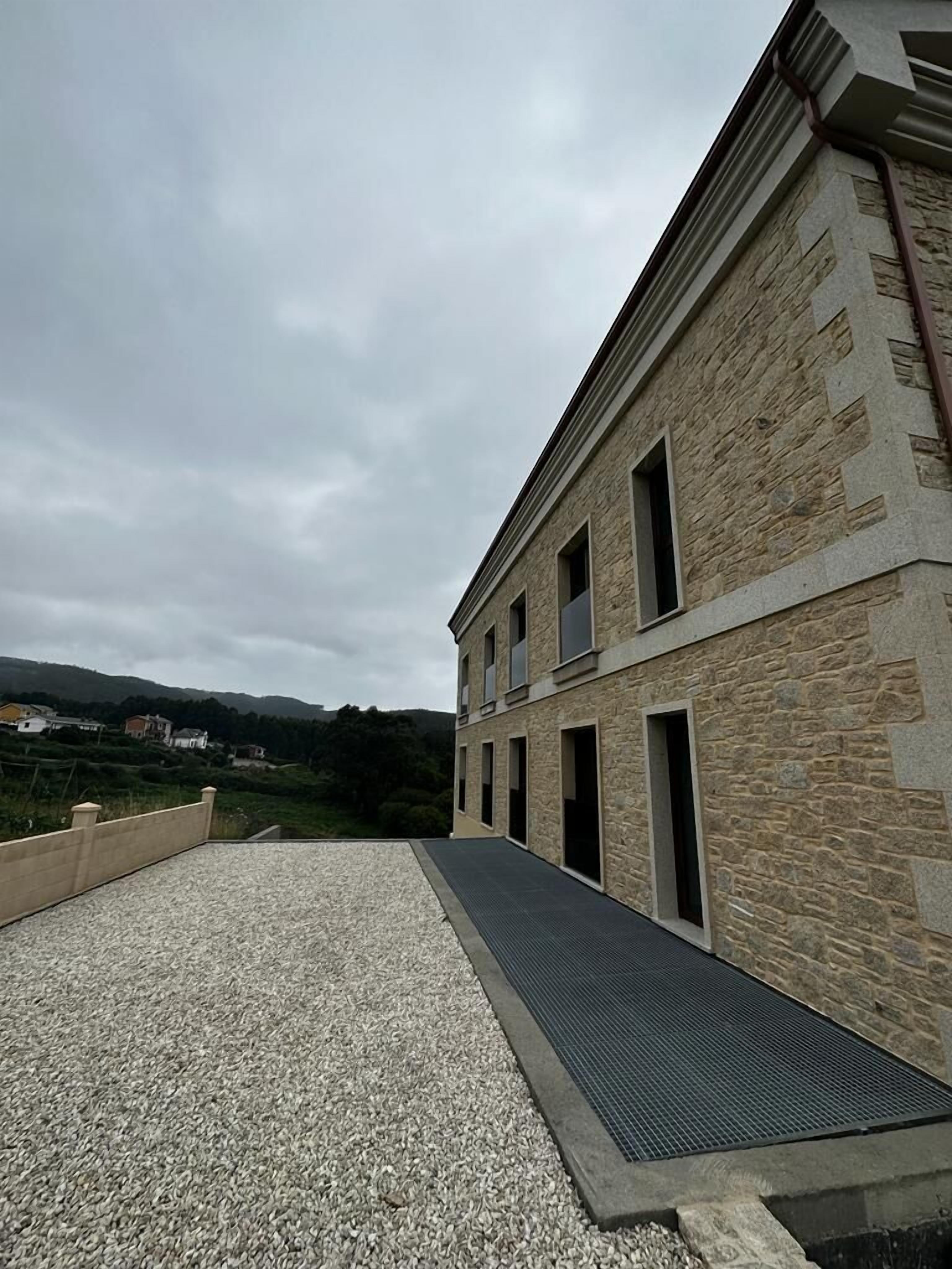 A stone building with multiple windows and a gravel pathway leading to a metal ramp.