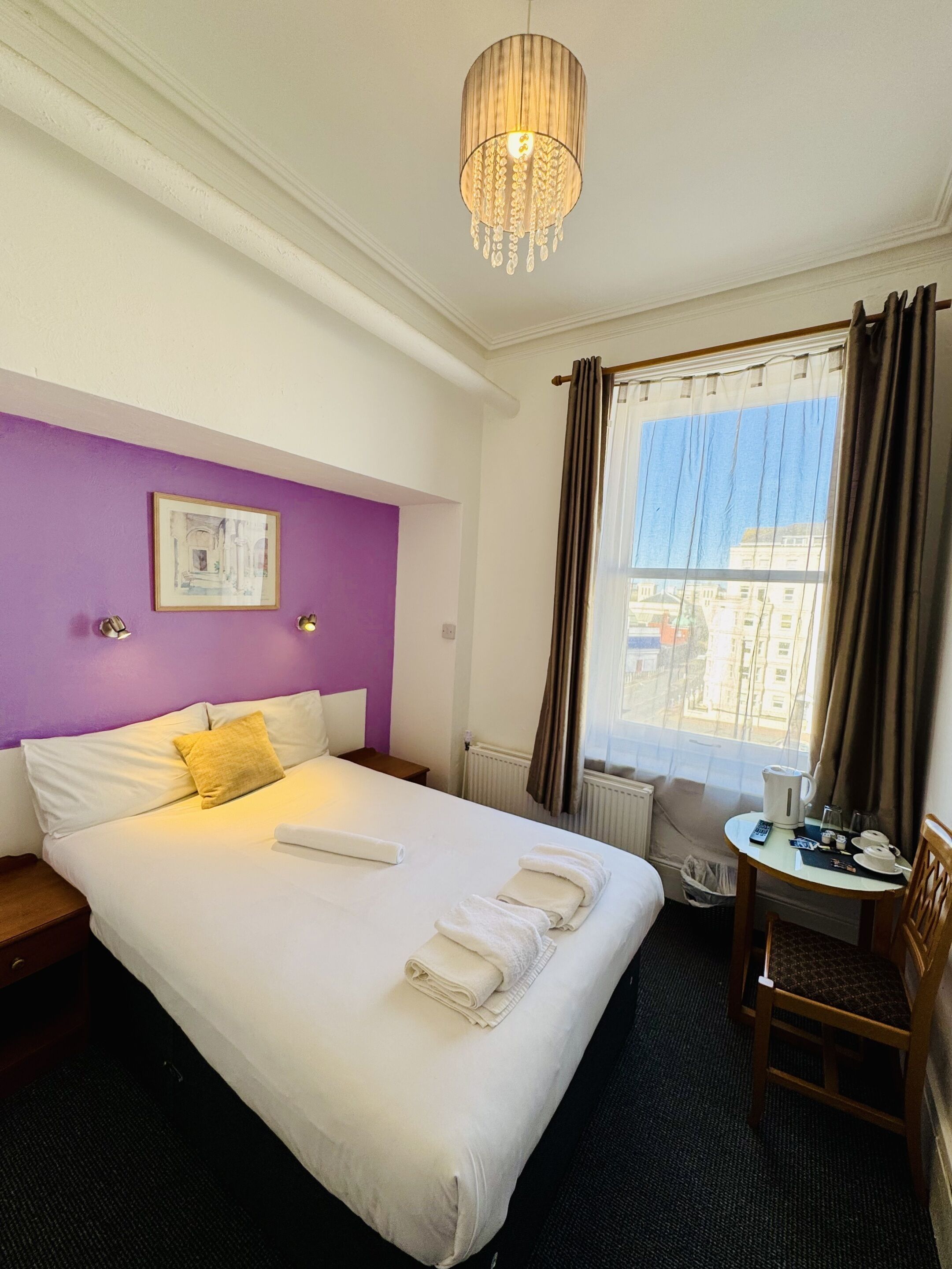 A hotel room with a large bed, a small table with a kettle and cups, a chair, a window with curtains, and a chandelier.