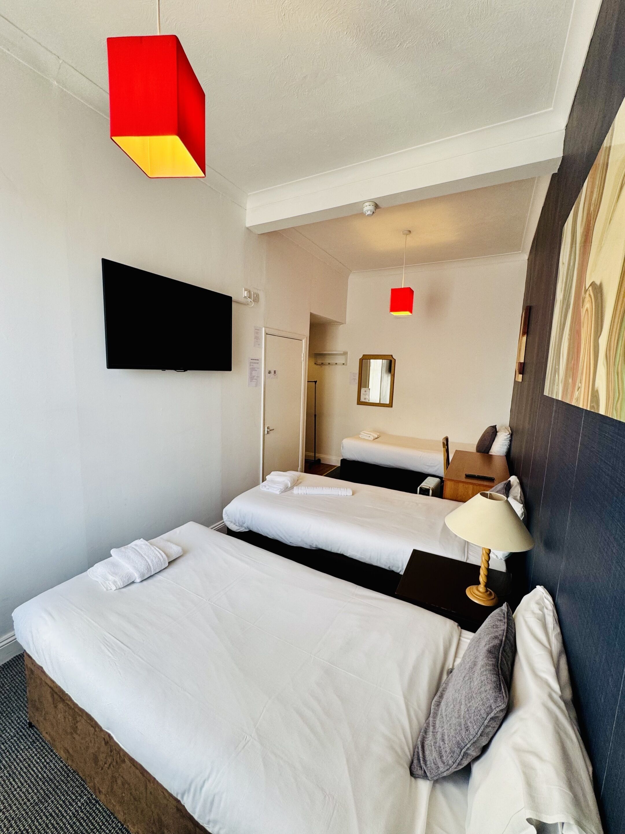 A hotel room with two beds, a flat-screen TV, a red pendant light, and a mirror on the wall.