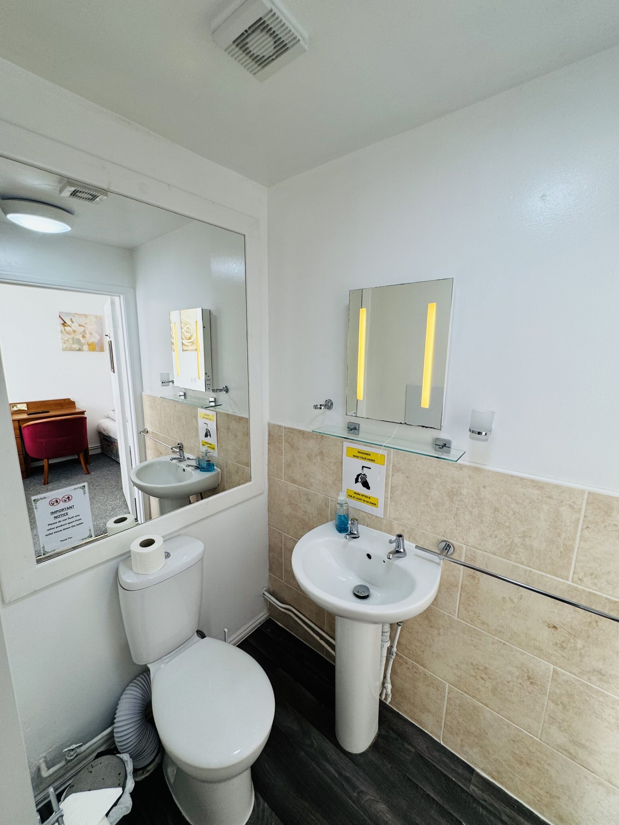 A bathroom with a toilet, sink, and mirror. There is a sign on the wall and a picture of a room in the mirror.