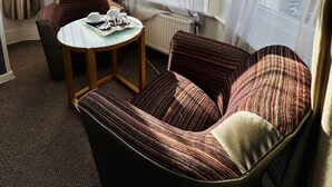 A cozy room with a round table, two striped armchairs, and a view of buildings through large windows.