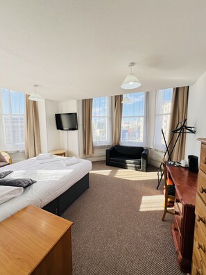A hotel room with a bed, a desk, a chair, a TV, a sofa, and large windows with curtains. - Carlton Courth Hotel Eastbourne (Eastbourne)