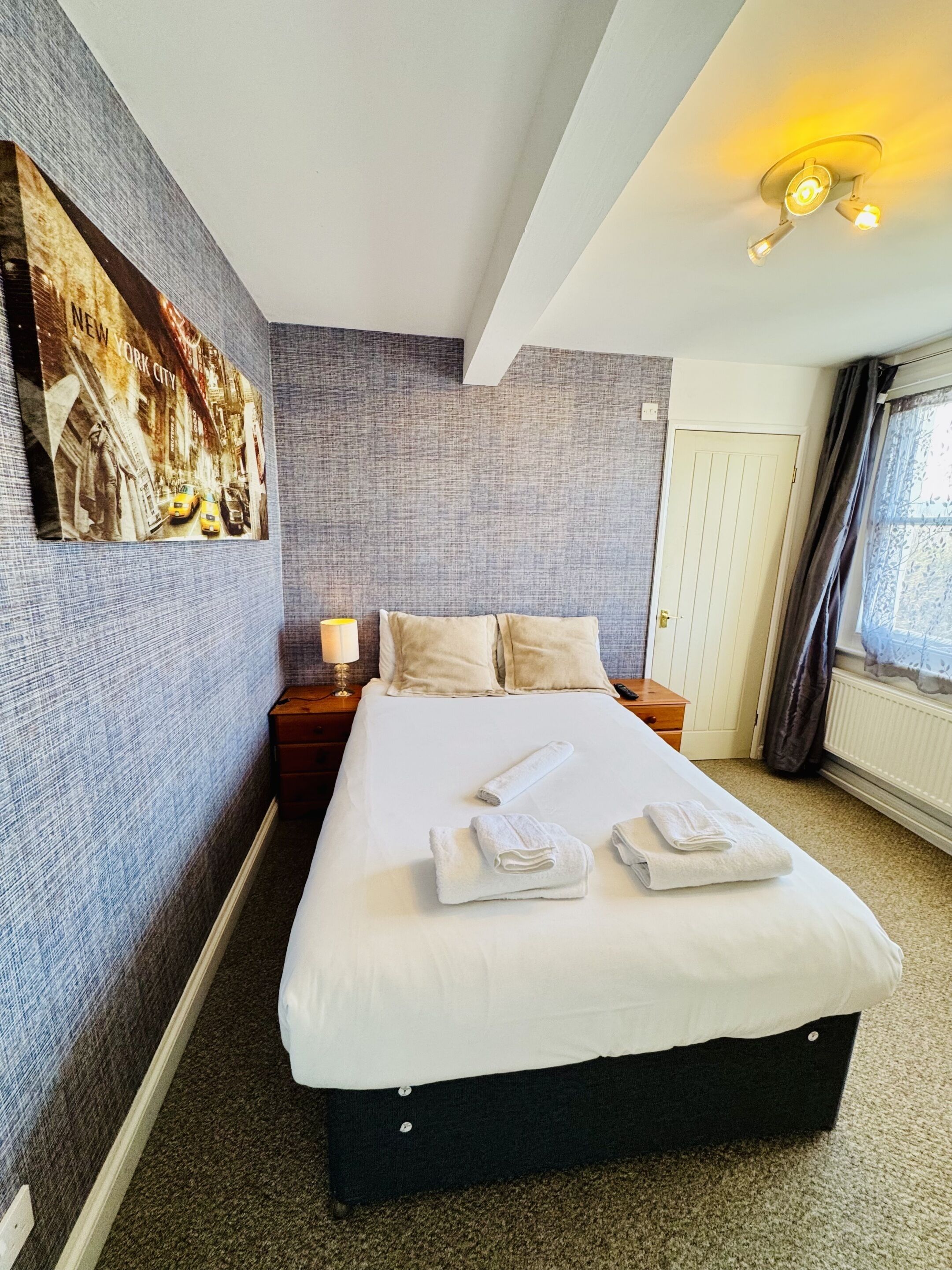 A hotel room with a large bed, a nightstand, a lamp, a painting on the wall, and a window with curtains.