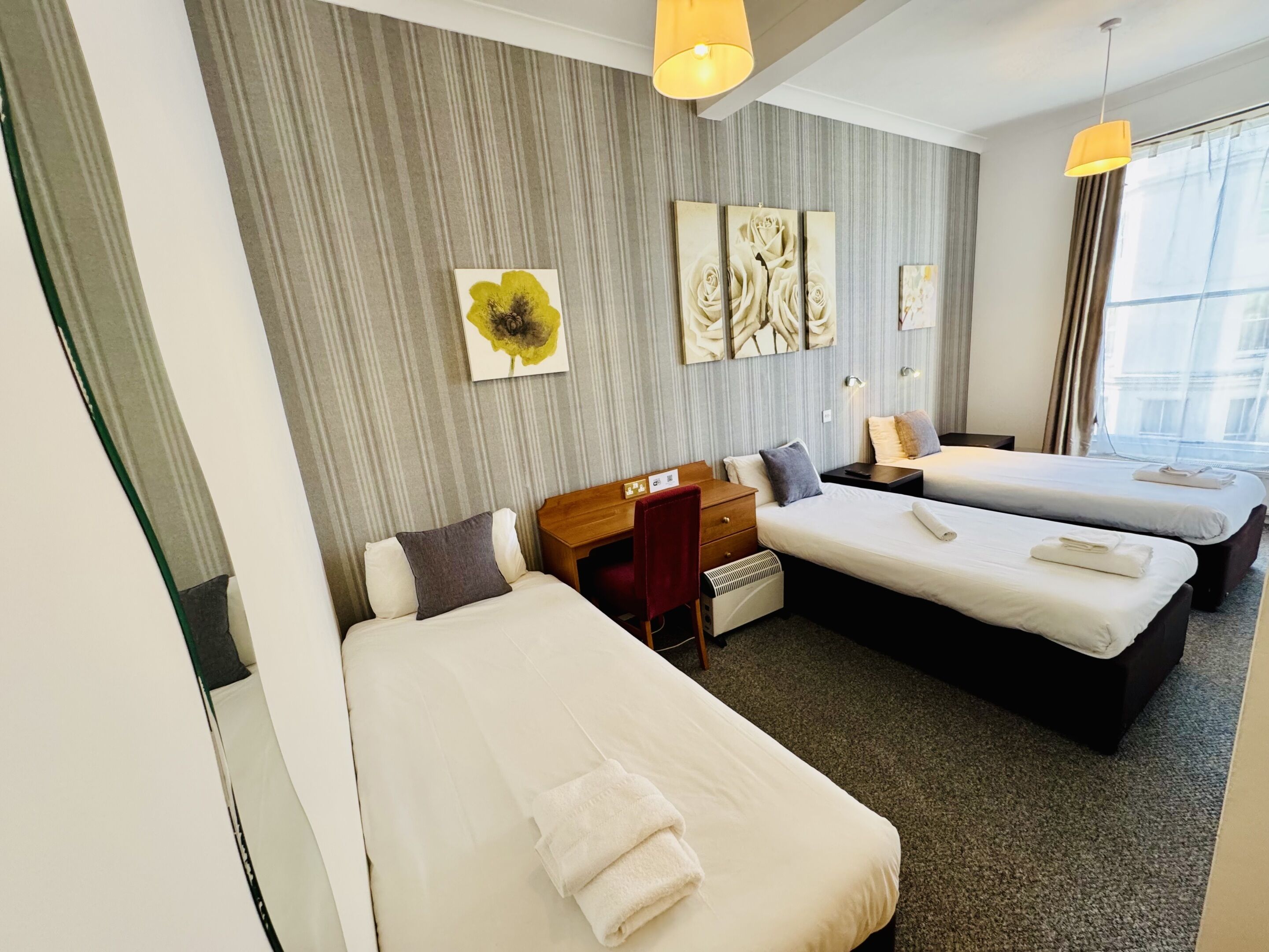 A hotel room with two beds, a desk, and a chair. There are wall art pieces and a window with curtains.