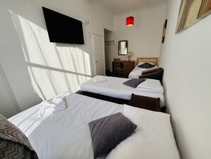 A hotel room with two beds, a desk, a chair, a TV, and a window with sunlight. - Carlton Courth Hotel Eastbourne (Eastbourne)
