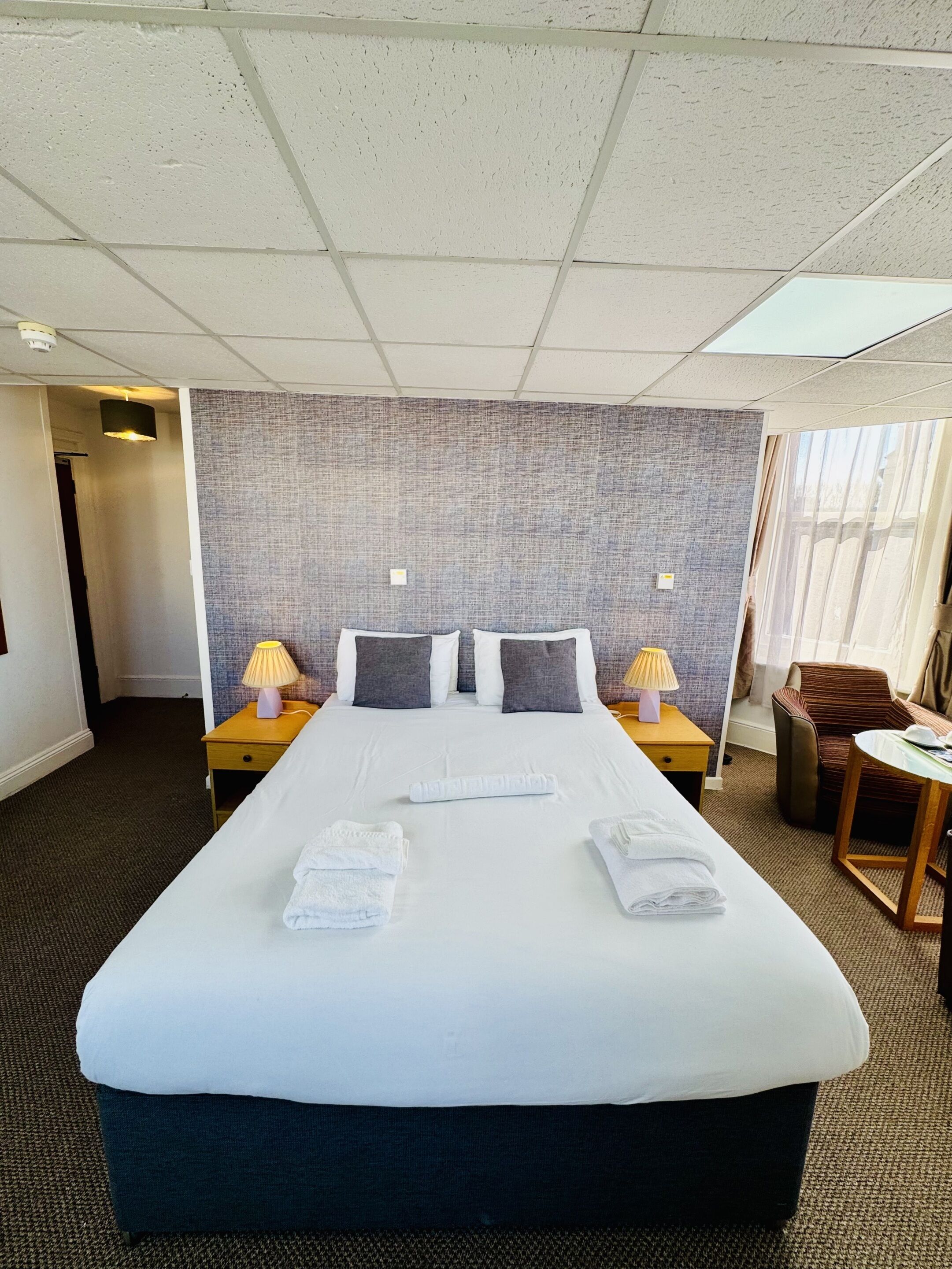 A hotel room with a large bed, two bedside lamps, a chair, and a small table.