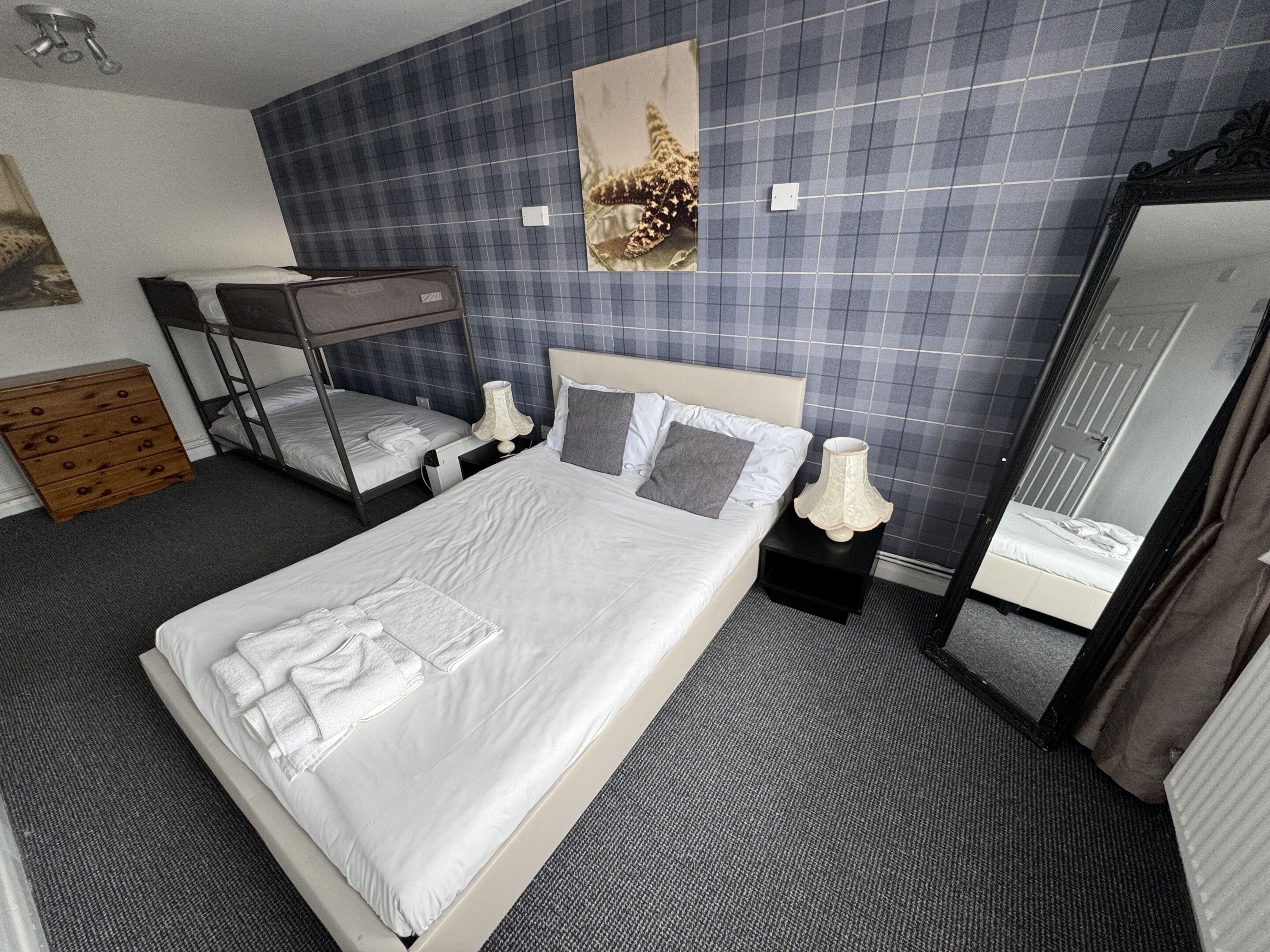 Quarto de hotel com cama grande, beliche, cómoda de madeira, espelho e parede com padrão xadrez.