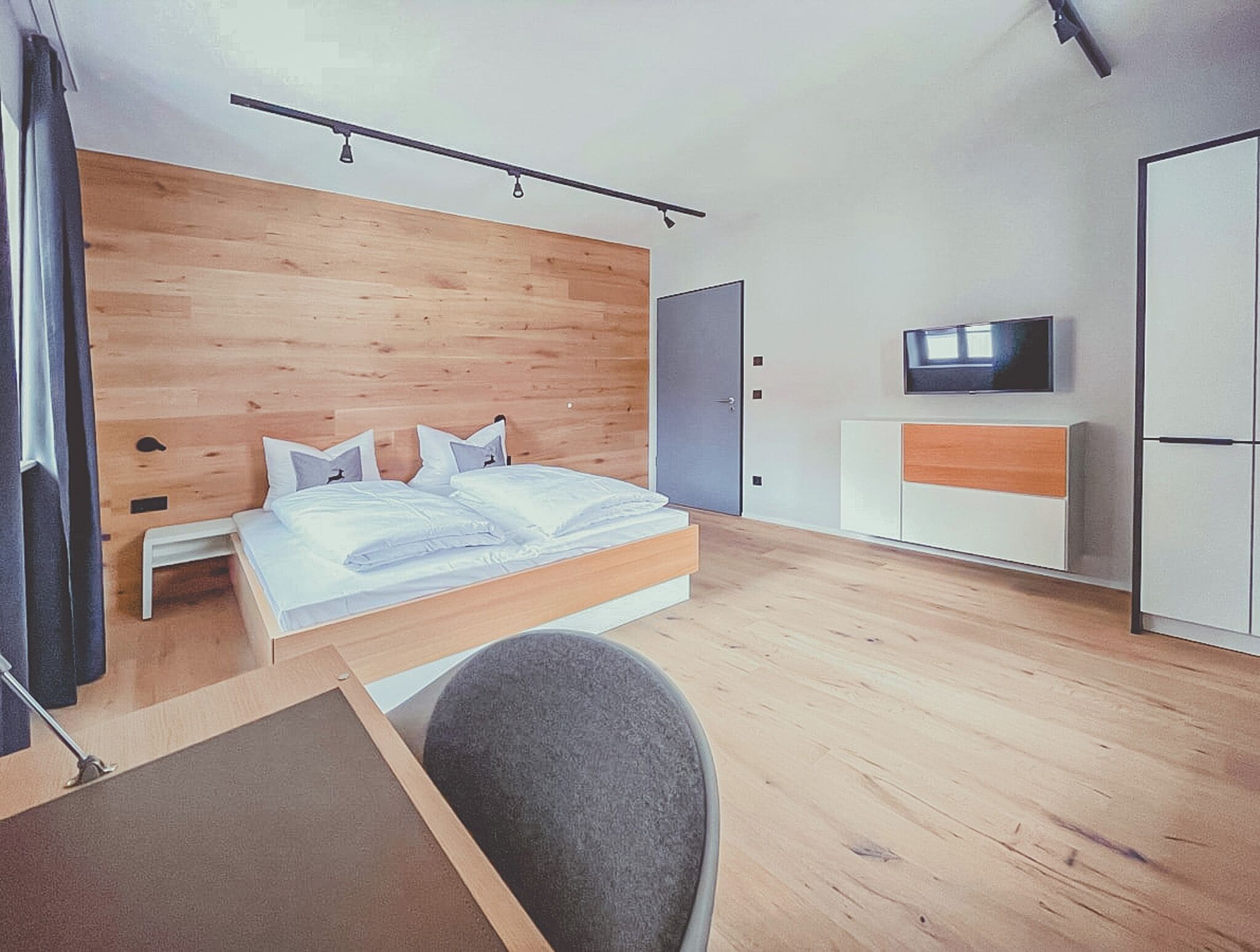 A modern bedroom with a wooden headboard, a bed with white bedding, a TV on the wall, and a wardrobe.