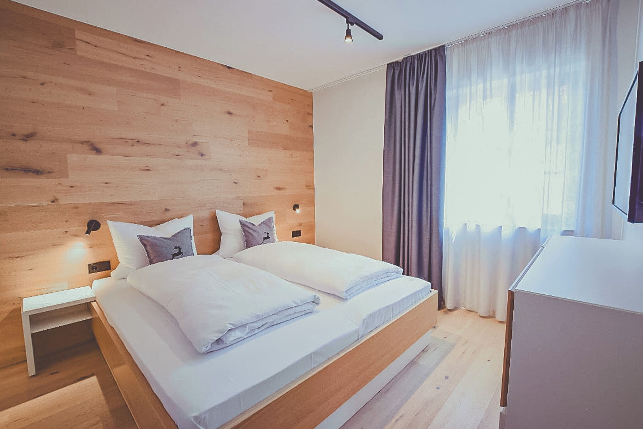 A modern bedroom with a wooden headboard, a large bed with white linens, a nightstand, and a desk with a chair.