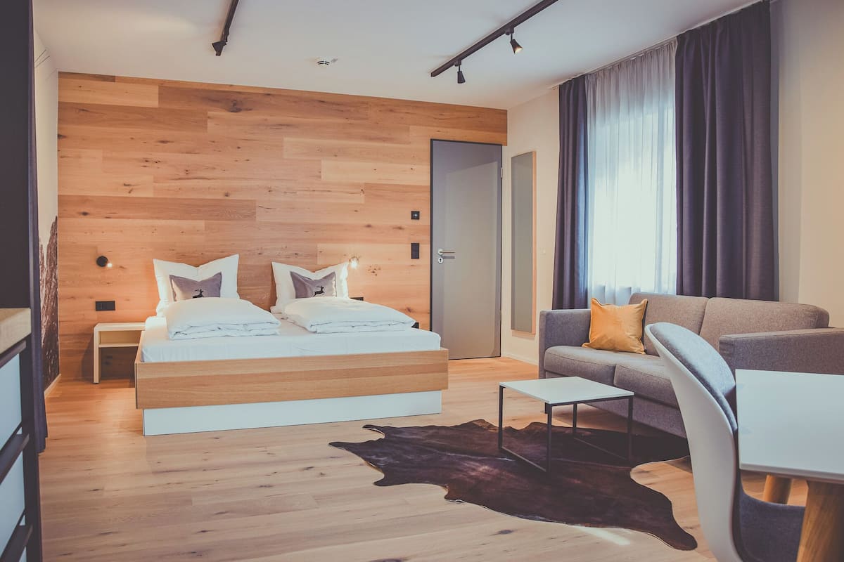Un dormitorio moderno con cabecera de madera, cama con ropa de cama blanca, sofá gris, una mesita y alfombra de cuero.