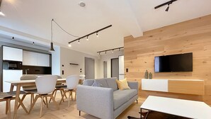 Ein modernes Wohnzimmer mit grauem Sofa, Holzboden, einem Essbereich mit Stühlen, einer Küche im Hintergrund und einem an der Wand befestigten Fernseher.