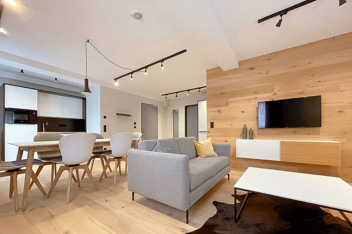 Una sala moderna con un sofá gris, piso de madera, un comedor con sillas, una cocina al fondo y un televisor montado en la pared.