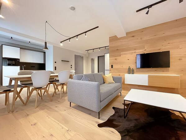 Una sala moderna con un sofá gris, piso de madera, un comedor con sillas, una cocina al fondo y un televisor montado en la pared.