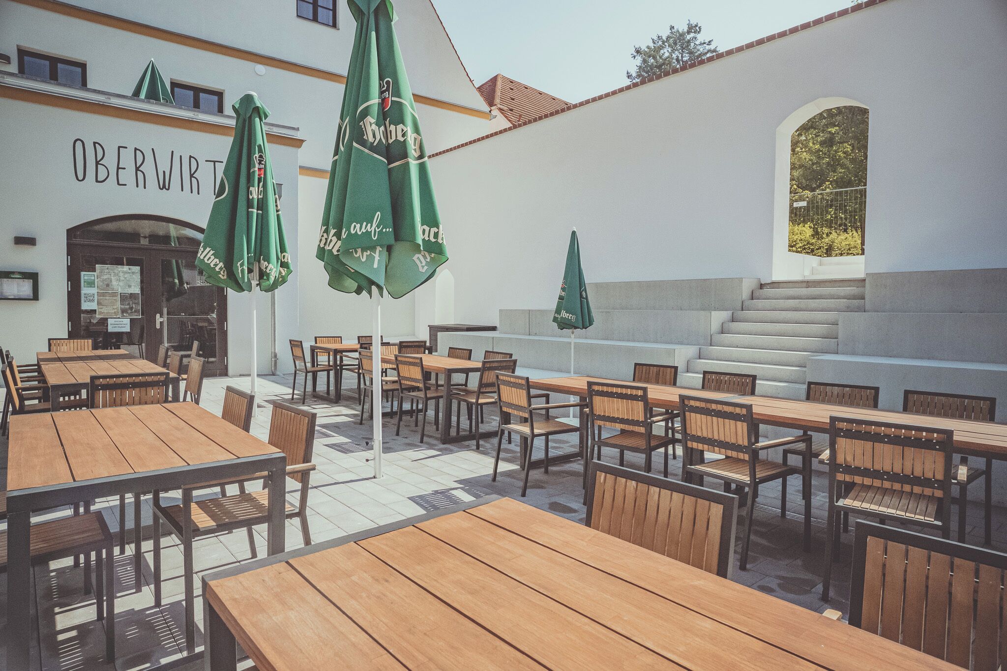 An outdoor seating area with wooden tables and chairs, green umbrellas, and a building with the sign "OBERWIRT".