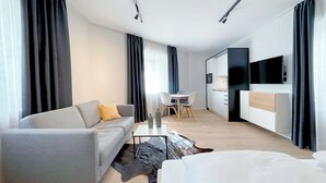 Ein modernes Hotelzimmer mit grauem Sofa, einem kleinen Esstisch, einem Flachbildfernseher und einem großen Fenster mit Vorhängen.