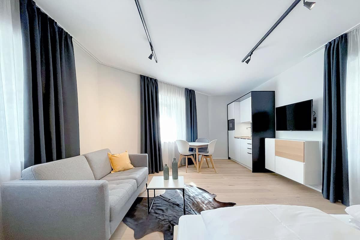 Una habitación de hotel moderna con un sofá gris, una pequeña mesa de comedor, un televisor de pantalla plana y un ventanal con cortinas.