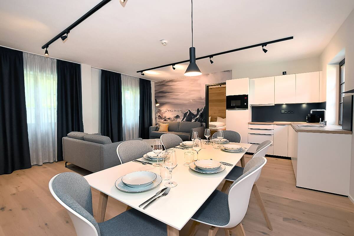 Un comedor moderno con una mesa larga dispuesta para seis comensales, rodeada de sillas tapizadas en gris. Se aprecia una cocina con armarios blancos y una encimera negra al fondo.