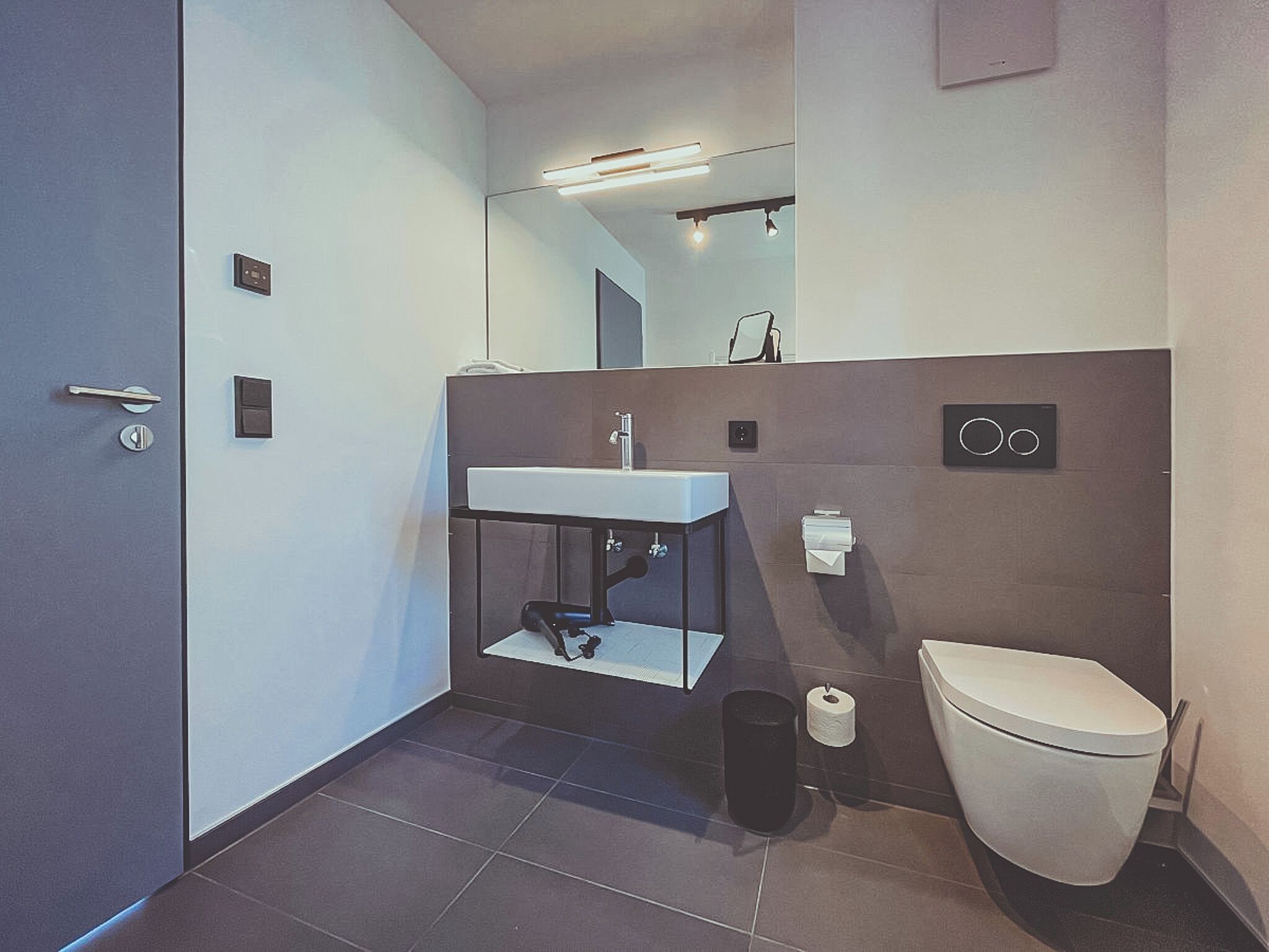 A modern bathroom with a sink, mirror, toilet, and a trash bin.
