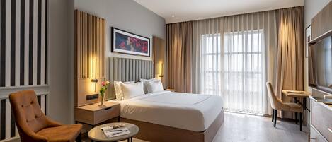 A modern hotel room with a large bed, a sitting area with a chair and a table, a TV, and a large window with curtains.