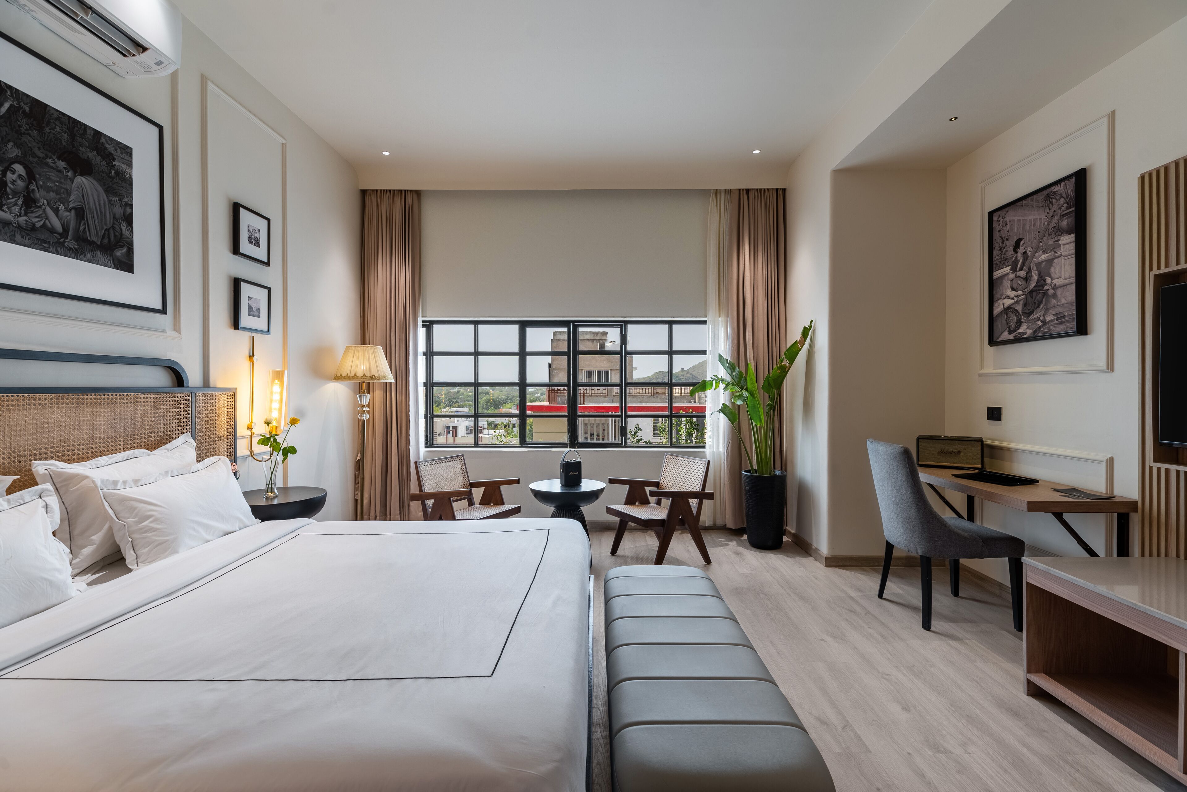 A hotel room with a large bed, a desk, a chair, a small table, and a potted plant.