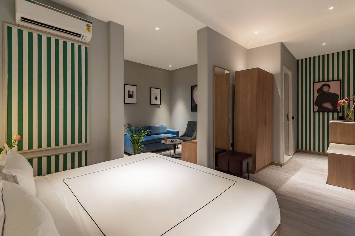 Una habitación de hotel moderna con una cama grande, un área de descanso con sofá y sillones, una pequeña mesa y un armario de madera.