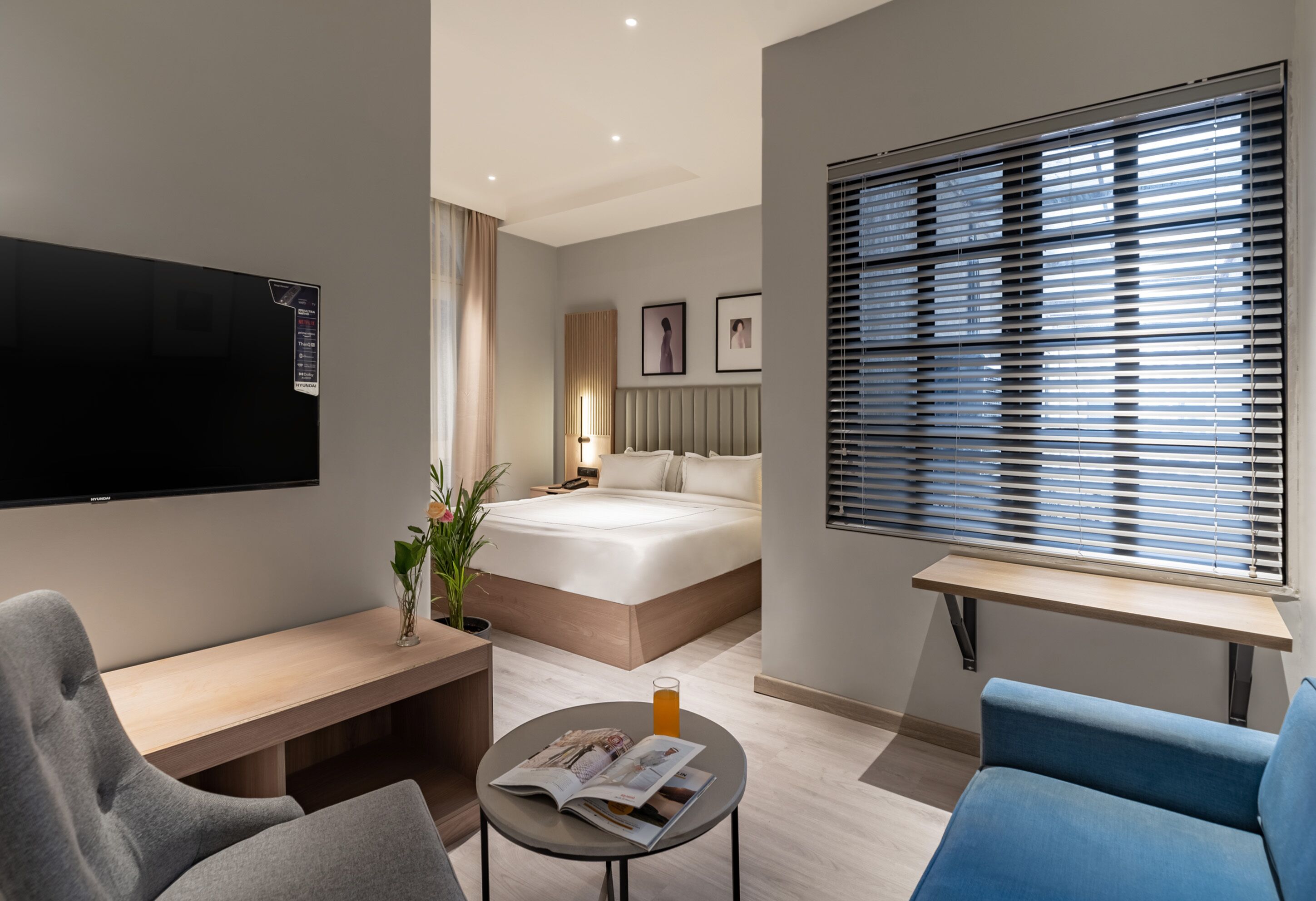 Une chambre d’hôtel moderne équipée d’un lit, d’une télévision, d’un bureau, d’une chaise, d’une petite table avec un magazine et d’une fenêtre avec des stores.