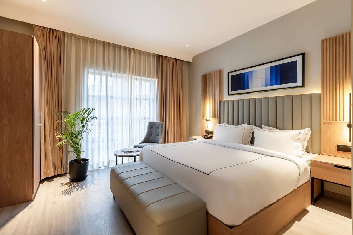 Una habitación de hotel moderna con una cama grande, una mesita de noche, una silla, una pequeña mesa y una planta en maceta.