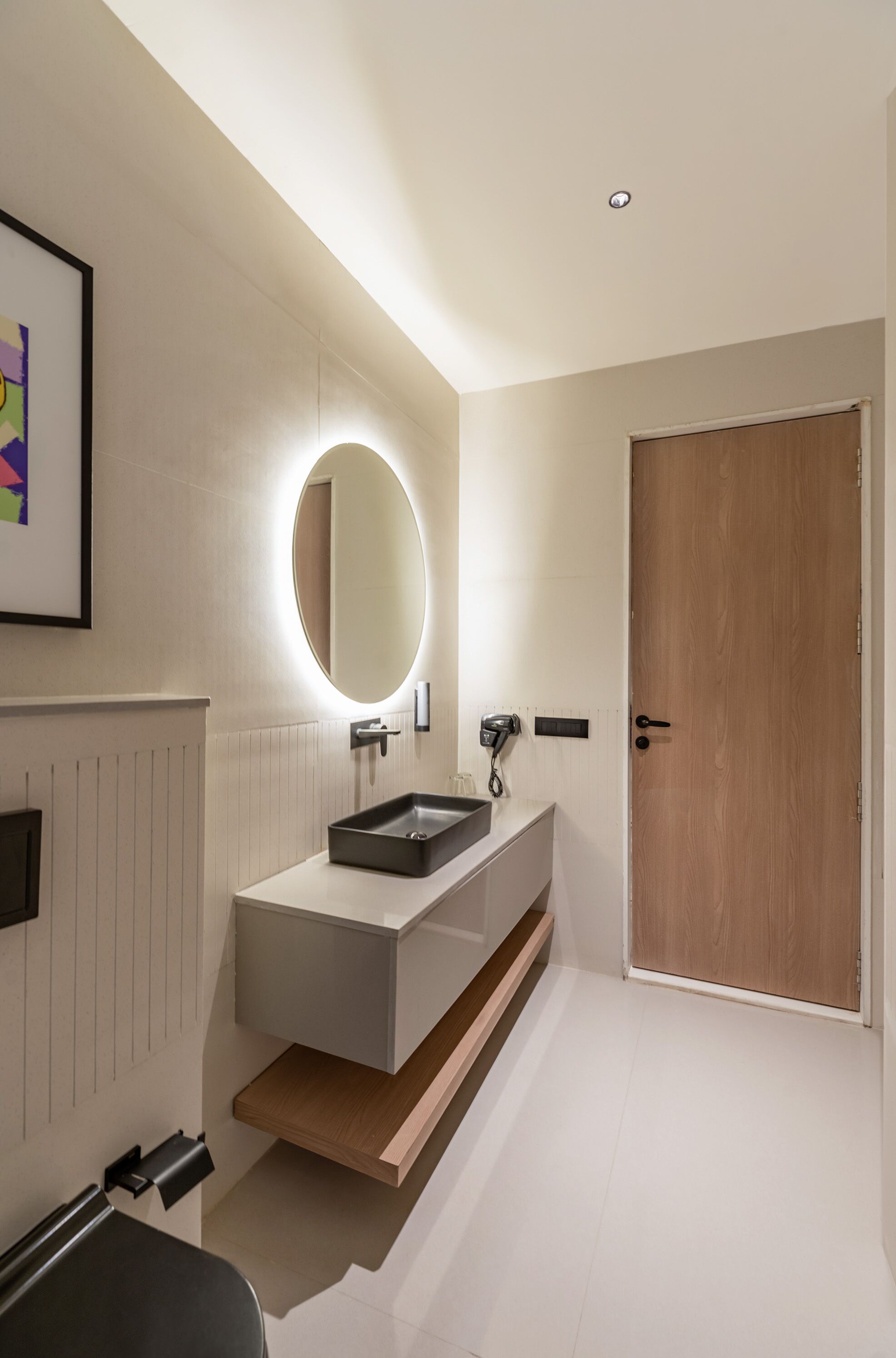 Un baño moderno con tocador blanco, un espejo redondo con iluminación, una puerta de madera y un cuadro en la pared.