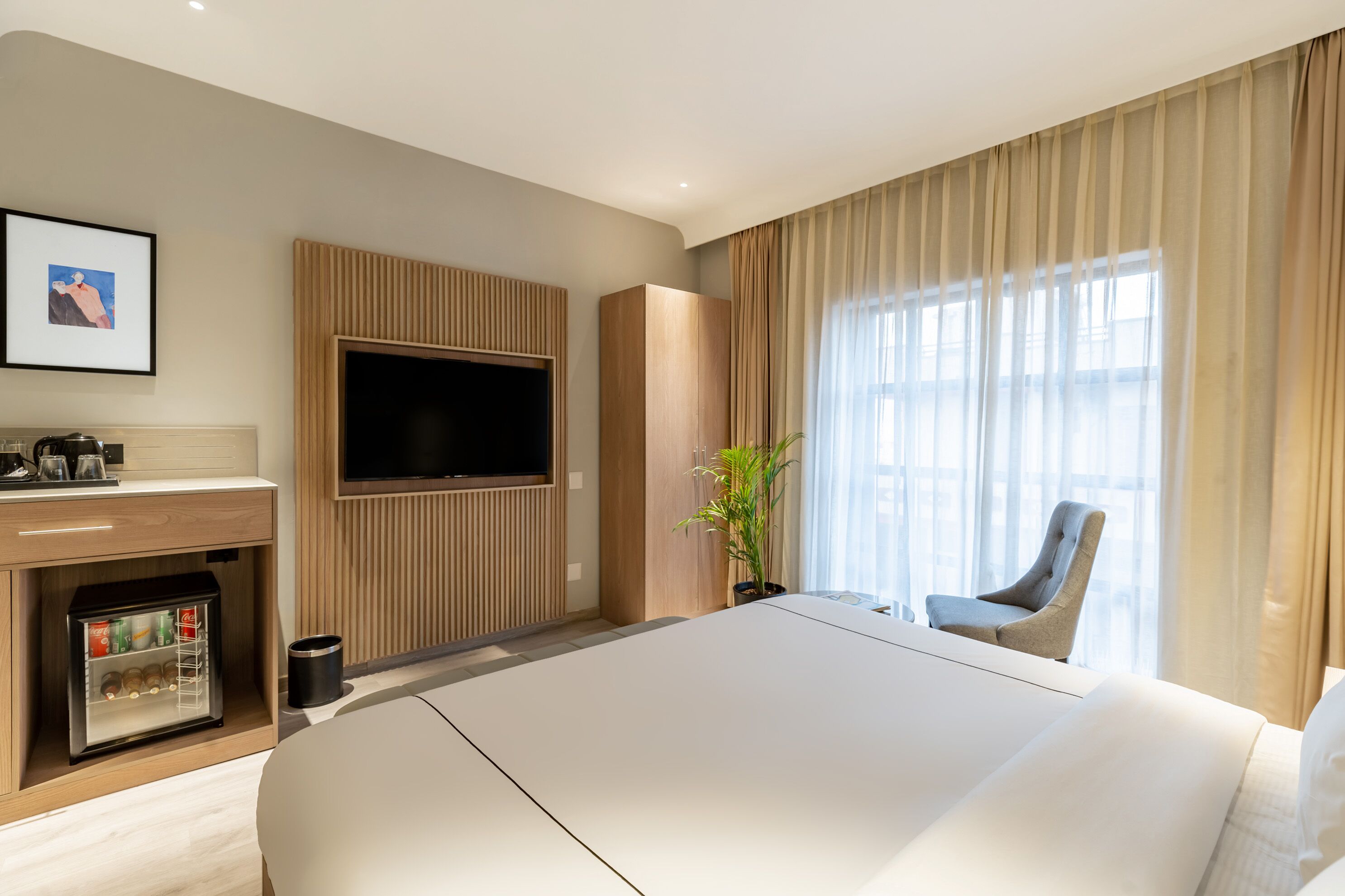Une chambre d’hôtel dotée d’un grand lit, d’un téléviseur à écran plat fixé au mur, d’un minibar, d’une plante en pot et offrant une vue sur la ville grâce à de grandes fenêtres.