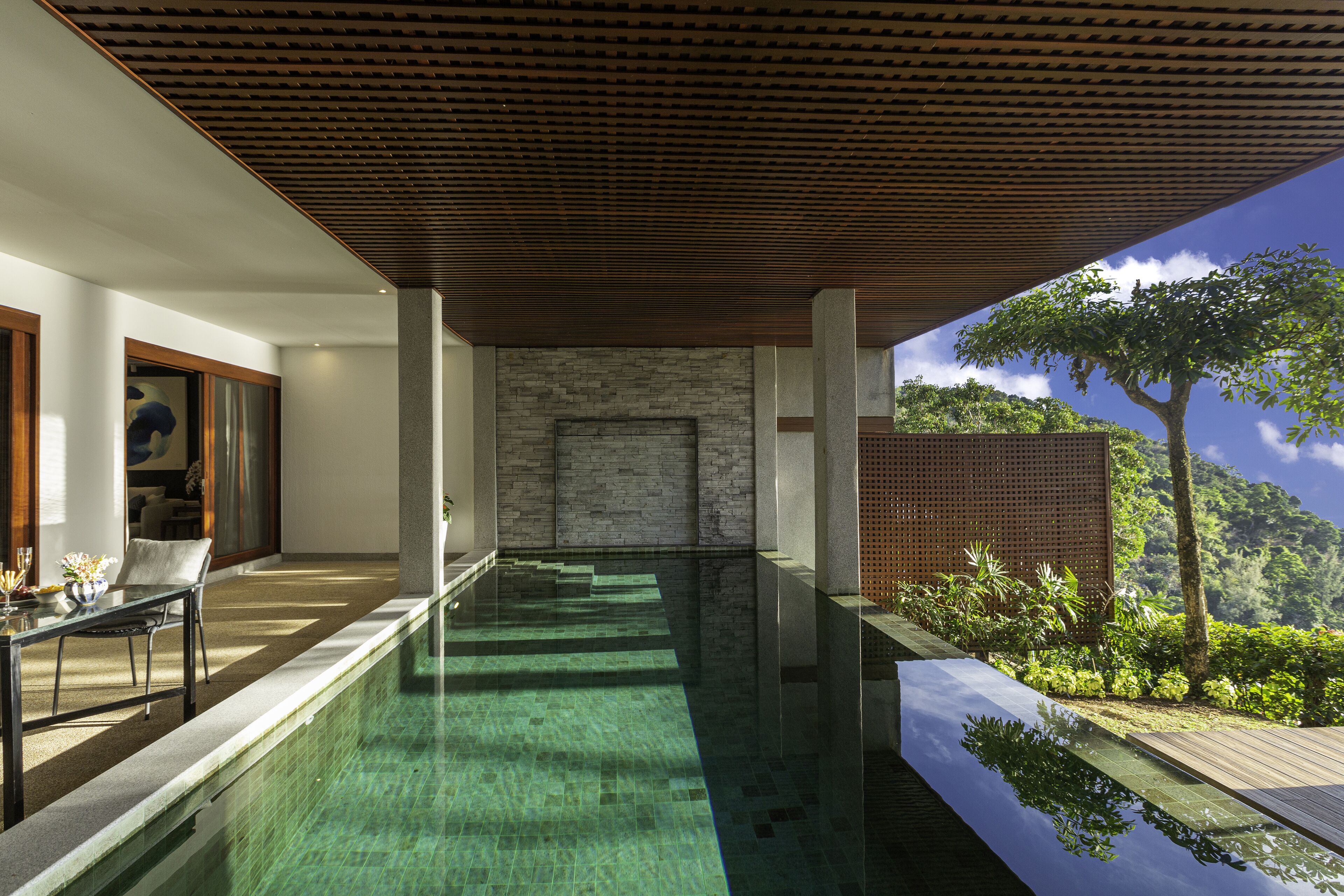 A modern indoor pool area with a clear view of the surrounding landscape, a stone wall, and a wooden ceiling.