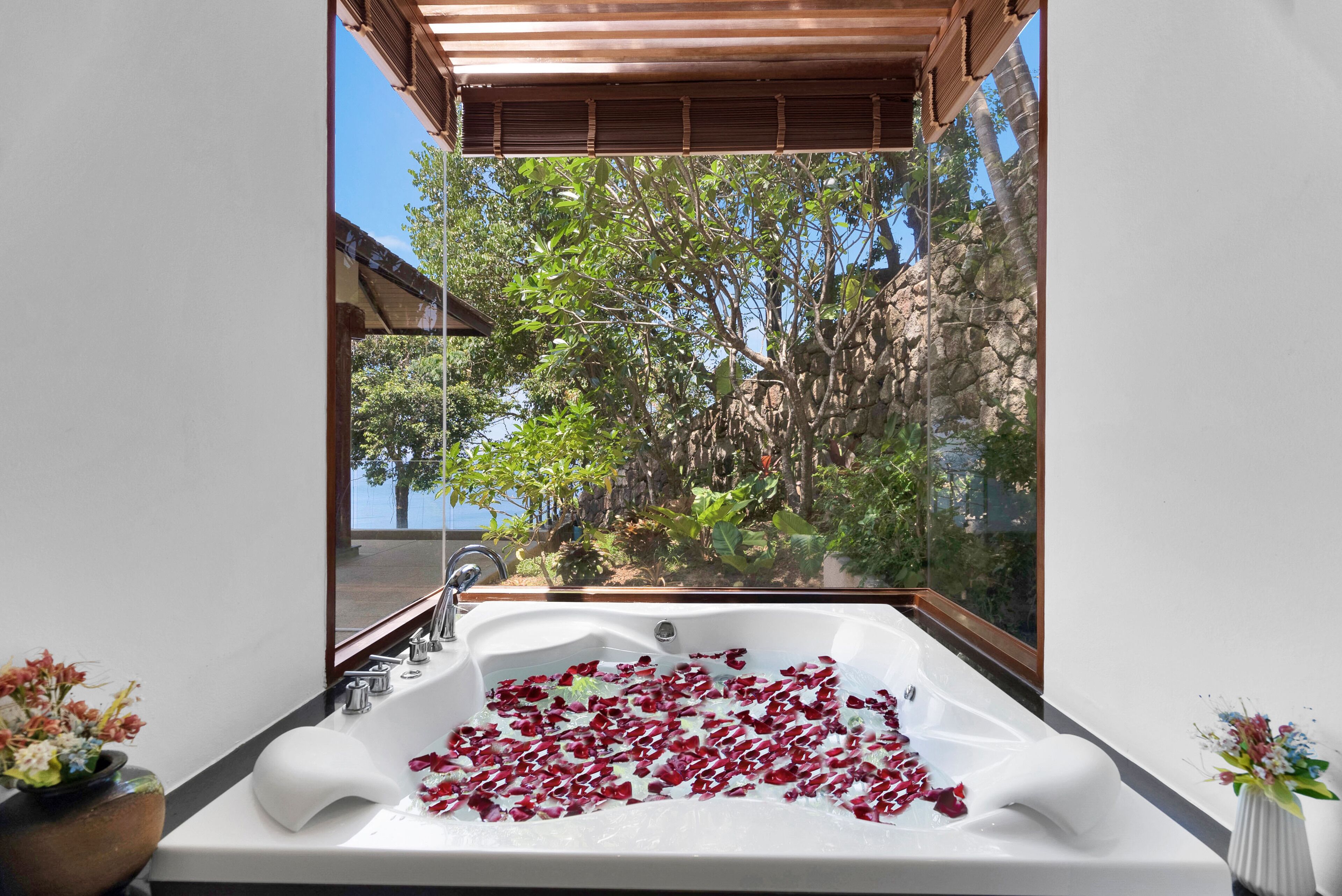 Un baño con una bañera grande llena de pétalos de rosa, techo de madera y vistas a árboles y un edificio a través de un ventanal.