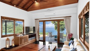 A spacious living room with a wooden ceiling, large windows offering a view of the ocean, a sofa with cushions, a coffee table, and a TV on a wooden cabinet. - Villa Varya (Kamala)
