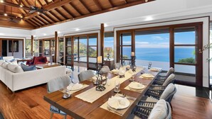 A spacious dining area with a long wooden table set for six, large windows offering a view of the ocean, and a high ceiling with exposed beams. - Villa Varya (Kamala)