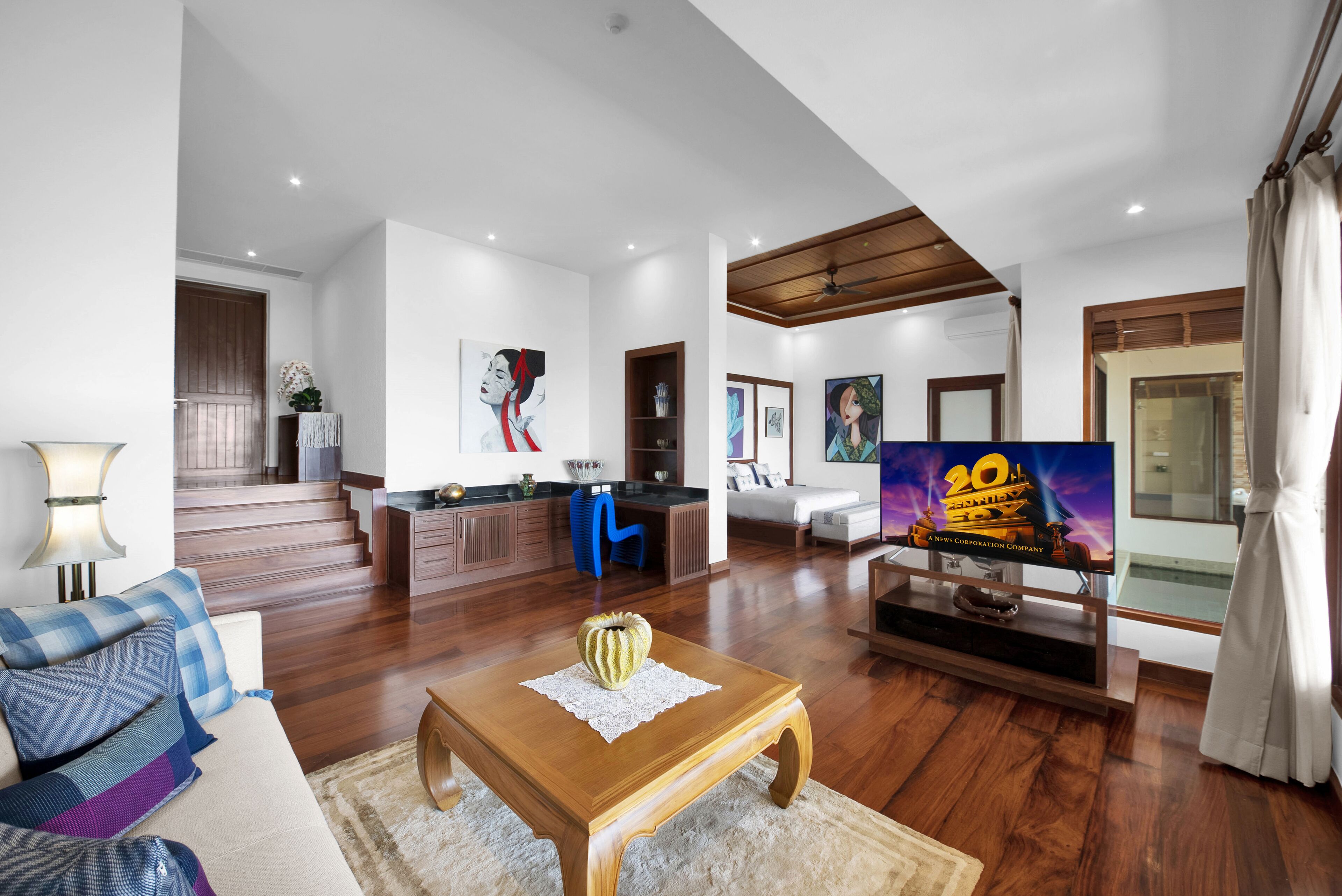A spacious living area with a wooden coffee table, a sofa with patterned cushions, a television, and a staircase leading to a kitchen area.