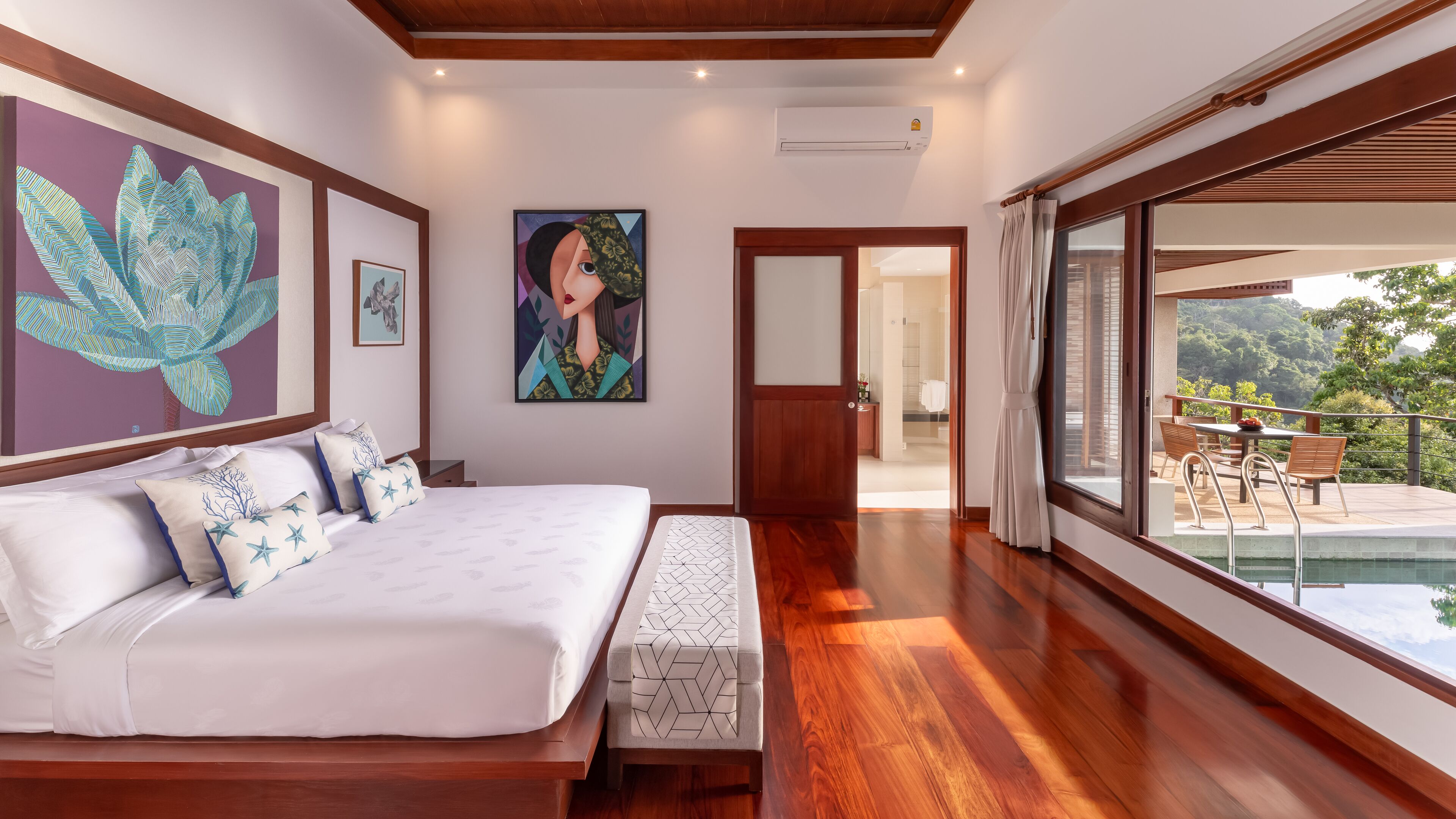 A bedroom with a large bed, a painting of a lotus flower, and a view of a balcony with outdoor furniture.
