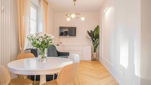 En moderne stue med et rundt spisebord, stole, et fladskærms-tv, en stor potteplante og en minimalistisk lampe.