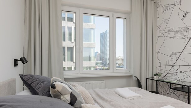 Un moderno cuarto de hotel con una cama grande, un escritorio con una silla, y vistas de la ciudad a través de amplias ventanas.