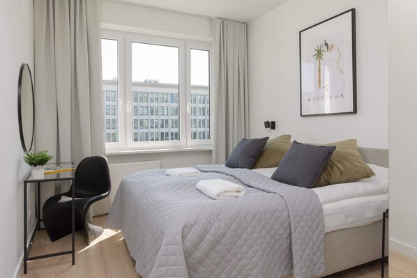 Ein modernes Schlafzimmer mit Bett, Schreibtisch und Stuhl. Ein Fenster mit Vorhängen, ein gerahmtes Bild an der Wand und eine Heizung sind ebenfalls vorhanden.