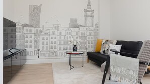 Una sala de estar moderna con un mural de paisaje urbano, un sofá negro, un sillón estampado y una pequeña mesa redonda con un florero.