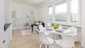 Un comedor moderno y bien iluminado con mobiliario blanco, un mural con un paisaje urbano en la pared y grandes ventanales con vistas a la ciudad.