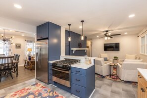 A modern kitchen with a built-in stove and oven, a dining area with a wooden table and chairs, and a living area with a sofa and a TV.