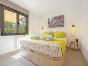 A modern bedroom with a large bed, a window with a view of trees, a cityscape painting, and a small table with a plant.