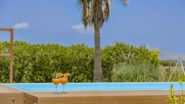 A wooden deck with two glasses of orange juice and a glass of water, overlooking a swimming pool and palm trees. - Villa Es Pont de Pedra (Alcúdia)