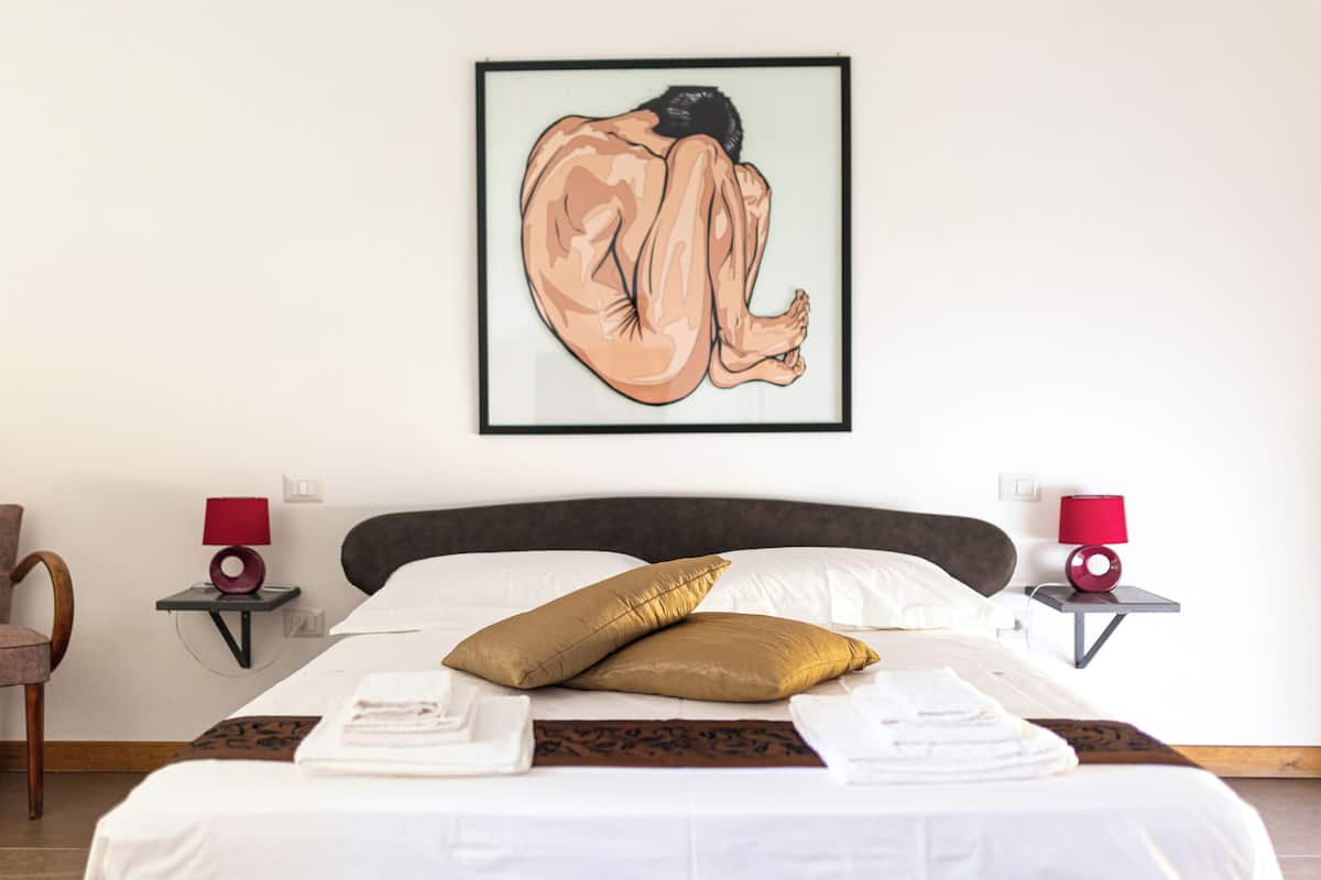Una cama bien hecha con sábanas blancas y un cojín dorado, dos mesitas de noche con lámparas rojas y una obra enmarcada en la pared.